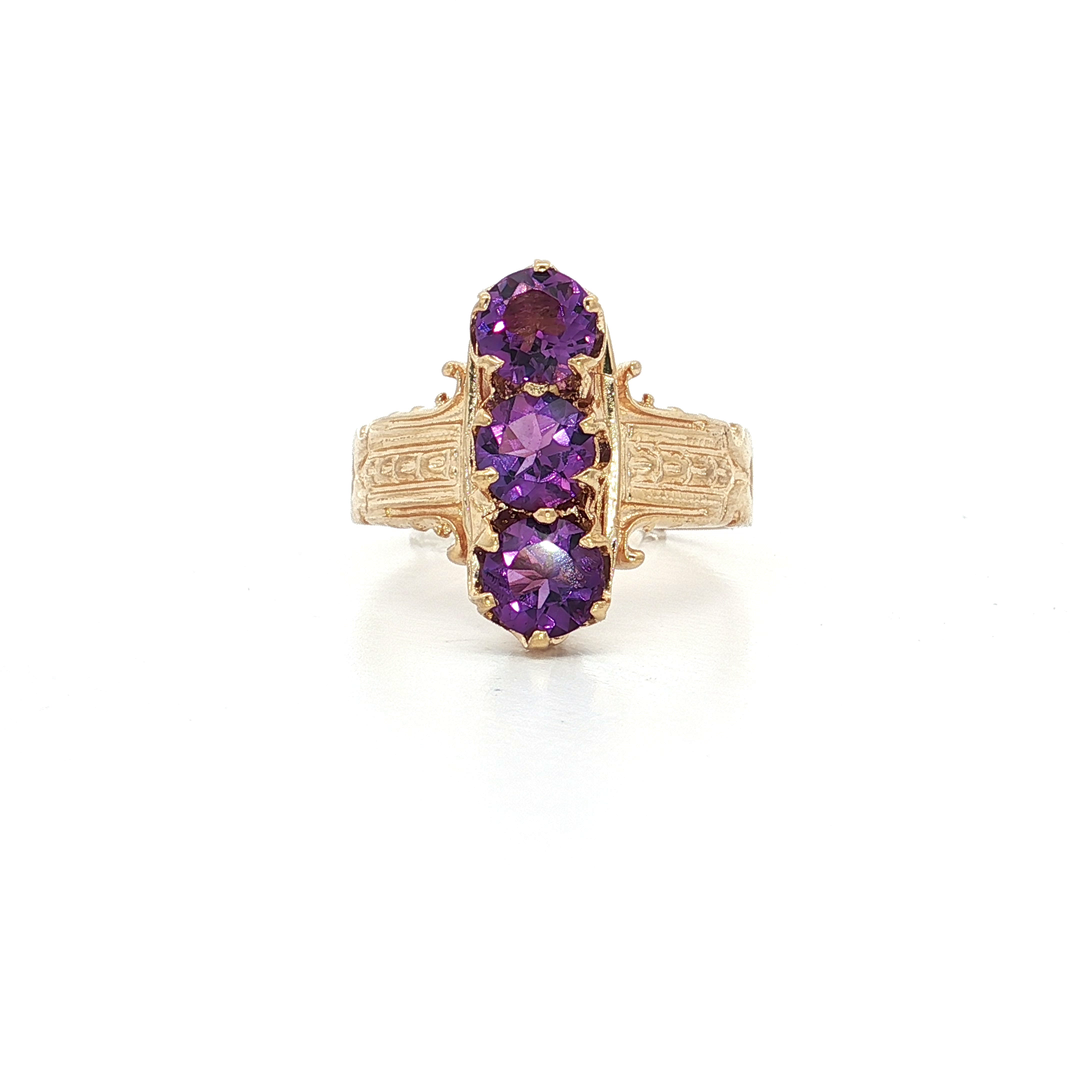 Vintage gold ring with three oval-cut purple gemstones, ornate band, white background