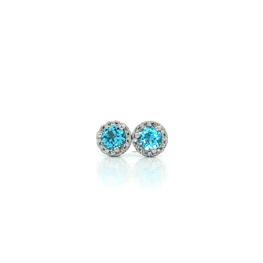 A pair of round stud earrings featuring bright blue gemstones surrounded by small clear stones in a silver-toned setting, displayed on a clean white background with a soft reflection.